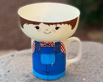 May include: A white ceramic bowl and mug set with a cartoon boy design. The boy is wearing blue overalls and a red and white checkered shirt. The bowl sits on top of the mug.