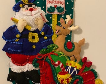 May include: A felt Christmas stocking with a Santa Claus dressed as a police officer writing a ticket to a reindeer. The stocking is decorated with sequins and has a green background with white polka dots. The sign says "No Parking".