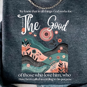May include: Dark gray sweatshirt with the text "The Good" and a colorful graphic. The sweatshirt features a gold chain necklace and the text "We know that in all things God works for..."