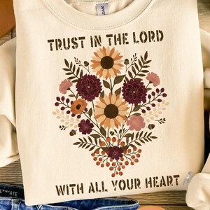 May include: Cream-colored sweatshirt with the text "TRUST IN THE LORD WITH ALL YOUR HEART" and a floral design. The floral design includes sunflowers, burgundy flowers, and other blooms. The sweatshirt is folded on a wooden surface.