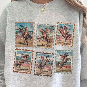Op de afbeelding: Een lichtgrijze sweatshirt met zes postzegelachtige afbeeldingen van rodeo cowboys op wilde paarden. De afbeeldingen zijn in bruin-, beige- en blauwtinten. De sweatshirt heeft lange mouwen en een ronde hals.