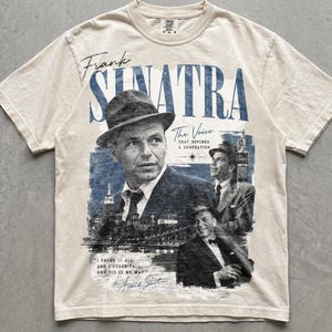 May include: An ivory short-sleeve t-shirt featuring a blue graphic print of Frank Sinatra. The design includes the text "Frank Sinatra," "The Voice That Defined A Generation," and the lyrics "I faced it all and I stood tall, and did it my way."