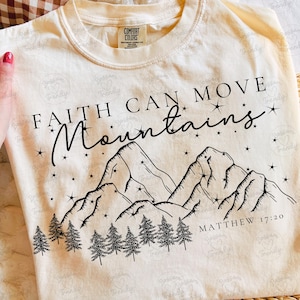 May include: Cream-colored t-shirt with the text "FAITH CAN MOVE Mountains" and a mountain graphic. The shirt also includes the Bible verse Matthew 17:20. The shirt is made of a soft material.
