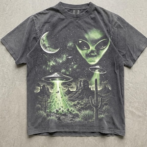 以下が含まれることがあります： チャコールグレーの半袖Tシャツ。緑色のエイリアンの顔、三日月、サボテンのある砂漠の風景の上空に浮かぶ2機の空飛ぶ円盤が描かれたグラフィックプリントが施されており、地面には光のビームが照射されている様子が表現されています。
