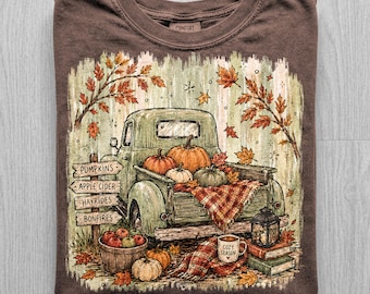 May include: A brown t-shirt featuring a graphic of a vintage green pickup truck loaded with pumpkins and a plaid blanket. The scene includes a wooden signpost labelled PUMPKINS, APPLE CIDER, HAYRIDES, BONFIRES, a basket of apples, a lantern, and a mug labelled COZY SEASON.