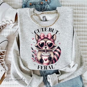 May include: Grey sweatshirt featuring a cute cartoon raccoon wearing pink sunglasses and a pink bow. The raccoon is holding a cup of coffee and the text "Cute But Feral" is printed on the sweatshirt.