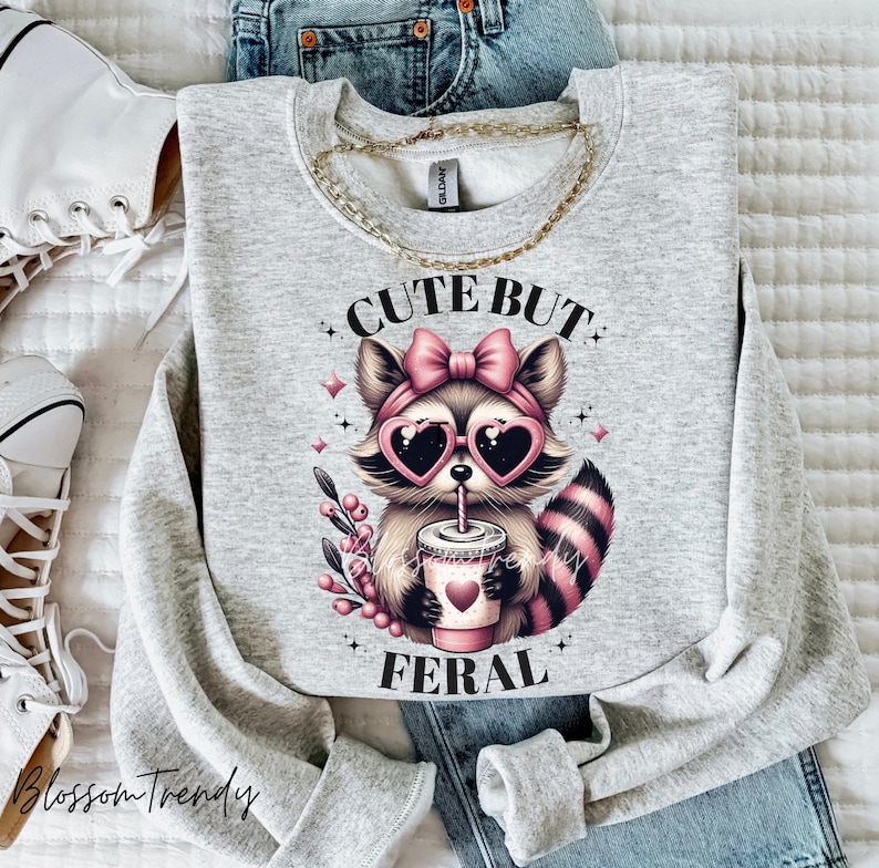 May include: Grey sweatshirt featuring a cute cartoon raccoon wearing pink sunglasses and a pink bow. The raccoon is holding a cup of coffee and the text "Cute But Feral" is printed on the sweatshirt.