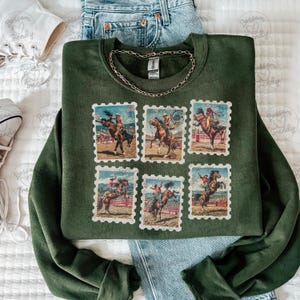 Op de afbeelding: Een donkergroene sweatshirt met zes postzegelontwerpen met rodeo-sc&egrave;nes. De postzegels tonen cowboys op wilde paarden. Een gouden ketting ligt op de sweatshirt. De sweatshirt wordt gecombineerd met een blauwe jeans en witte sneakers.