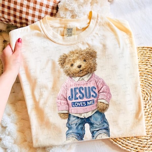Peut inclure: T-shirt cr&egrave;me avec une illustration d'ours en peluche. L'ours porte un pull rose avec les mots "JESUS LOVES ME" en bleu. L'ours porte &eacute;galement un jean bleu et des baskets blanches. Le t-shirt a une &eacute;tiquette Comfort Colors.