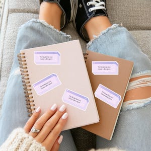 May include: A light pink spiral notebook and a brown notebook with several stickers that read "My brain has too many tabs open." The notebooks are on a person's lap, with their legs and feet in the background.