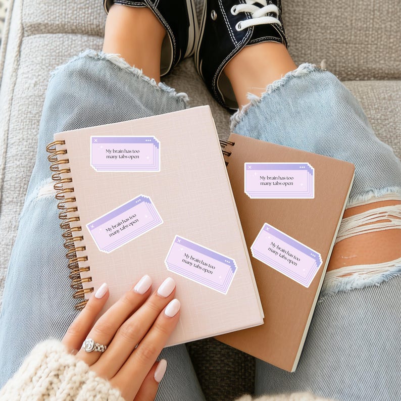 May include: A light pink spiral notebook and a brown notebook with several stickers that read "My brain has too many tabs open." The notebooks are on a person's lap, with their legs and feet in the background.