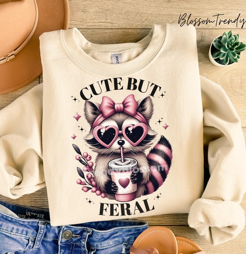May include: A cream-coloured sweatshirt featuring a cute cartoon raccoon wearing pink heart-shaped sunglasses and a pink bow. The raccoon is holding a pink cup with a straw and the text "Cute But Feral" is printed on the sweatshirt.