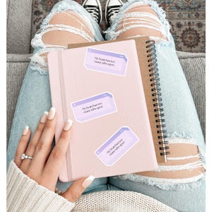 May include: A light pink journal and a brown spiral notebook are shown. Three stickers with the text "My brain has too many tabs open" are placed on the pink journal. The person is wearing ripped blue jeans and a white sweater.