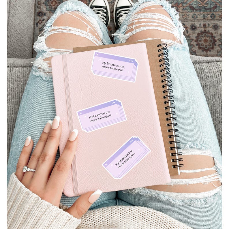 May include: A light pink journal and a brown spiral notebook are shown. Three stickers with the text "My brain has too many tabs open" are placed on the pink journal. The person is wearing ripped blue jeans and a white sweater.