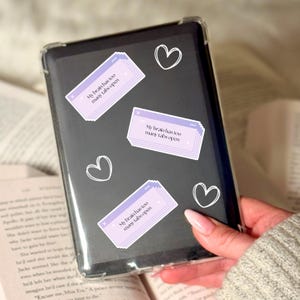 May include: A black tablet with a clear case is decorated with three light purple stickers that read "My brain has too many tabs open." White heart doodles are also on the tablet. The tablet is held over an open book.