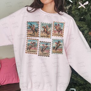 Op de afbeelding: Lichtroze sweatshirt met zes bruine postzegelontwerpen met rodeo cowboys op wilde paarden. De postzegels tonen cowboys in actie. De sweatshirt heeft een ronde hals en lange mouwen.