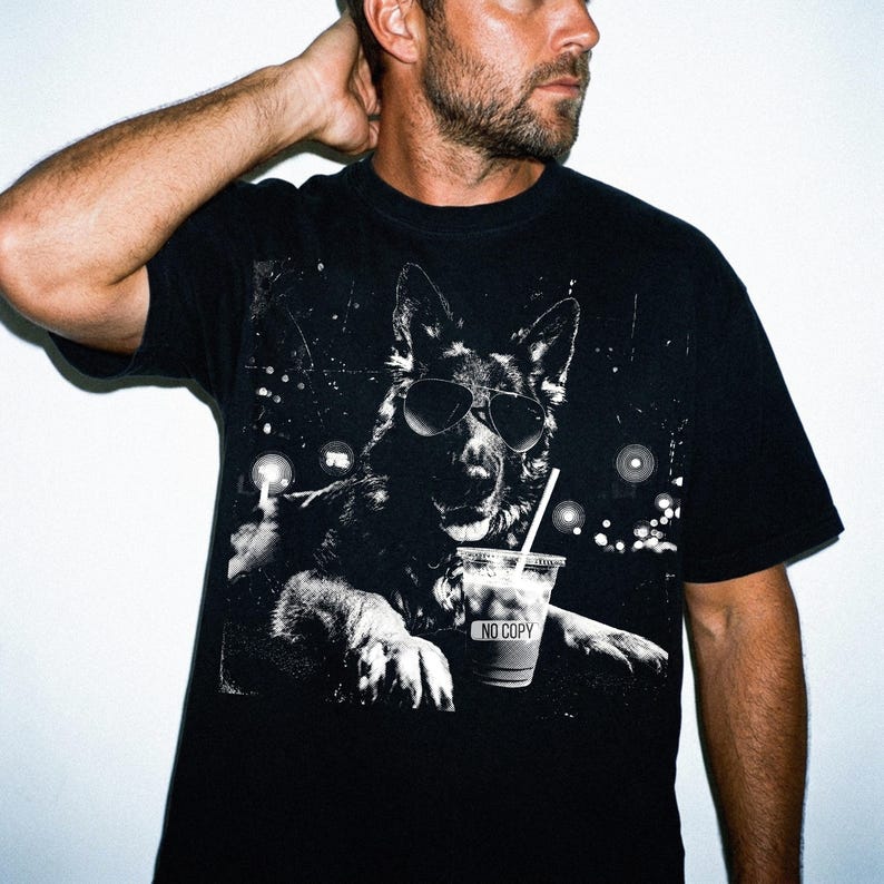 May include: Black t-shirt featuring a black and white graphic of a German Shepherd wearing sunglasses and holding a drink with a straw. The drink cup has the text "NO COPY". The dog is set against a background of lights.