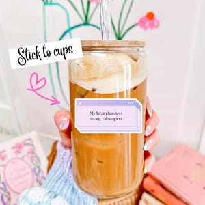 May include: A clear glass cup filled with a light brown beverage and topped with foam. The cup has a bamboo lid and a glass straw. Text on the cup reads "My brain has too many tabs open" and "Stick to cups".