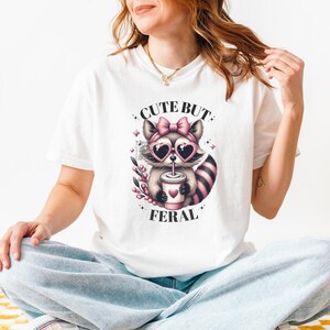 May include: White t-shirt featuring a graphic of a raccoon wearing pink sunglasses and a pink bow, drinking from a cup. The text "Cute But Feral" is printed on the shirt.