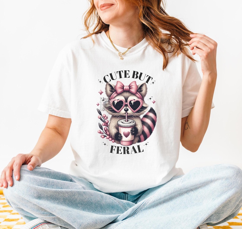 May include: White t-shirt featuring a graphic of a raccoon wearing pink sunglasses and a pink bow, drinking from a cup. The text "Cute But Feral" is printed on the shirt.