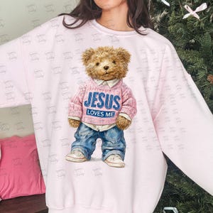 Peut inclure: Un sweat-shirt rose clair avec un ours en peluche portant un pull rose et un jean bleu. Le pull porte l'inscription "JESUS LOVES ME" en bleu et blanc. L'ours porte des baskets blanches.