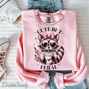 May include: Pink sweatshirt featuring a graphic of a raccoon wearing pink sunglasses and drinking from a cup. The text "Cute But Feral" is printed on the sweatshirt.