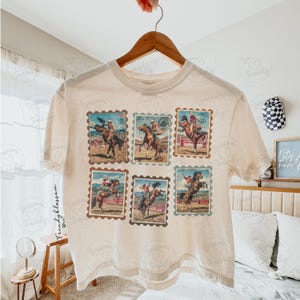 Op de afbeelding: Cr&egrave;mekleurig T-shirt met zes postzegelachtige afbeeldingen van rodeo cowboys die op bronco's rijden. De afbeeldingen zijn in bruin-, blauw- en roodtinten. Het shirt hangt aan een houten hanger.