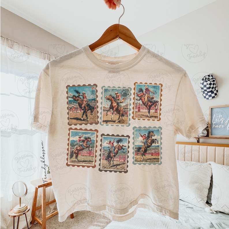 Op de afbeelding: Cr&egrave;mekleurig T-shirt met zes postzegelachtige afbeeldingen van rodeo cowboys die op bronco's rijden. De afbeeldingen zijn in bruin-, blauw- en roodtinten. Het shirt hangt aan een houten hanger.