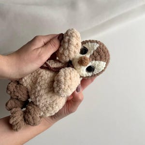 May include: A small, handmade crochet plush toy shaped like a sloth. The toy features a textured, cream-colored body with brown limbs and a brown and white face. It has large, round, black plastic eyes and a small brown nose, crafted from soft yarn.