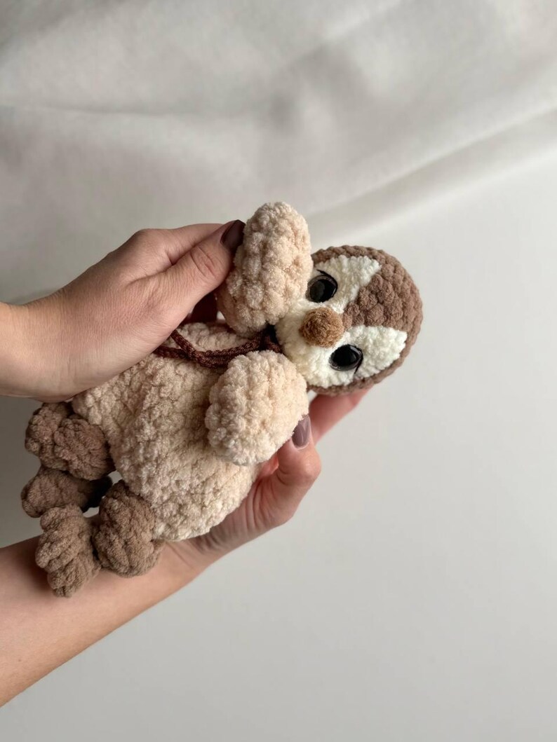 May include: A small, handmade crochet plush toy shaped like a sloth. The toy features a textured, cream-colored body with brown limbs and a brown and white face. It has large, round, black plastic eyes and a small brown nose, crafted from soft yarn.