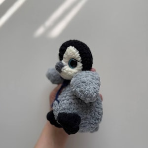 May include: A small, handmade crochet penguin plush toy featuring a textured grey body, a black head with a white face patch, large blue eyes, a small grey beak, and black feet. A thin dark blue ribbon is tied around the neck of the plush figure.
