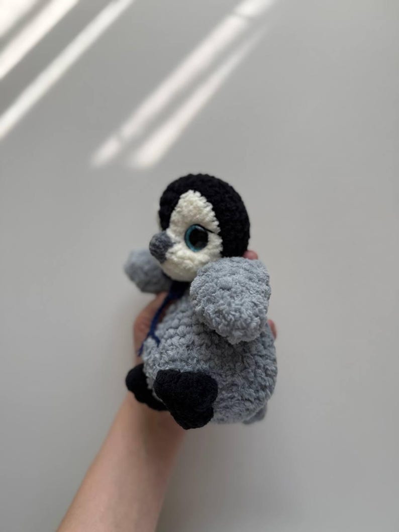 May include: A small, handmade crochet penguin plush toy featuring a textured grey body, a black head with a white face patch, large blue eyes, a small grey beak, and black feet. A thin dark blue ribbon is tied around the neck of the plush figure.