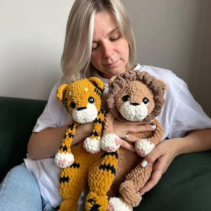 Può includere: Una donna tiene in mano due animali di peluche all'uncinetto. Uno è un leone marrone con una criniera soffice e l'altro è una tigre gialla e nera. Entrambi gli animali hanno zampe bianche e nasi rosa.