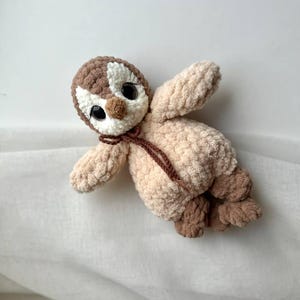 May include: A plush crochet toy shaped like a penguin with a brown and cream head, large black plastic eyes, and a light beige body. The toy features a small brown nose, a thin brown tie around the neck, and textured brown feet.