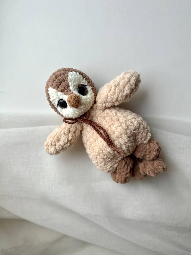 May include: A plush crochet toy shaped like a penguin with a brown and cream head, large black plastic eyes, and a light beige body. The toy features a small brown nose, a thin brown tie around the neck, and textured brown feet.