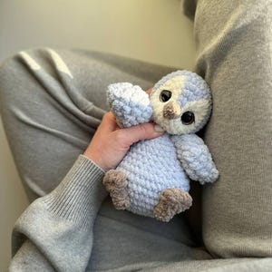 May include: A small, handmade crochet plush toy shaped like a penguin. The bird features a soft, light blue and cream-colored yarn body with a textured, fuzzy appearance. It has large, dark, glossy plastic eyes and a small, light brown yarn beak.