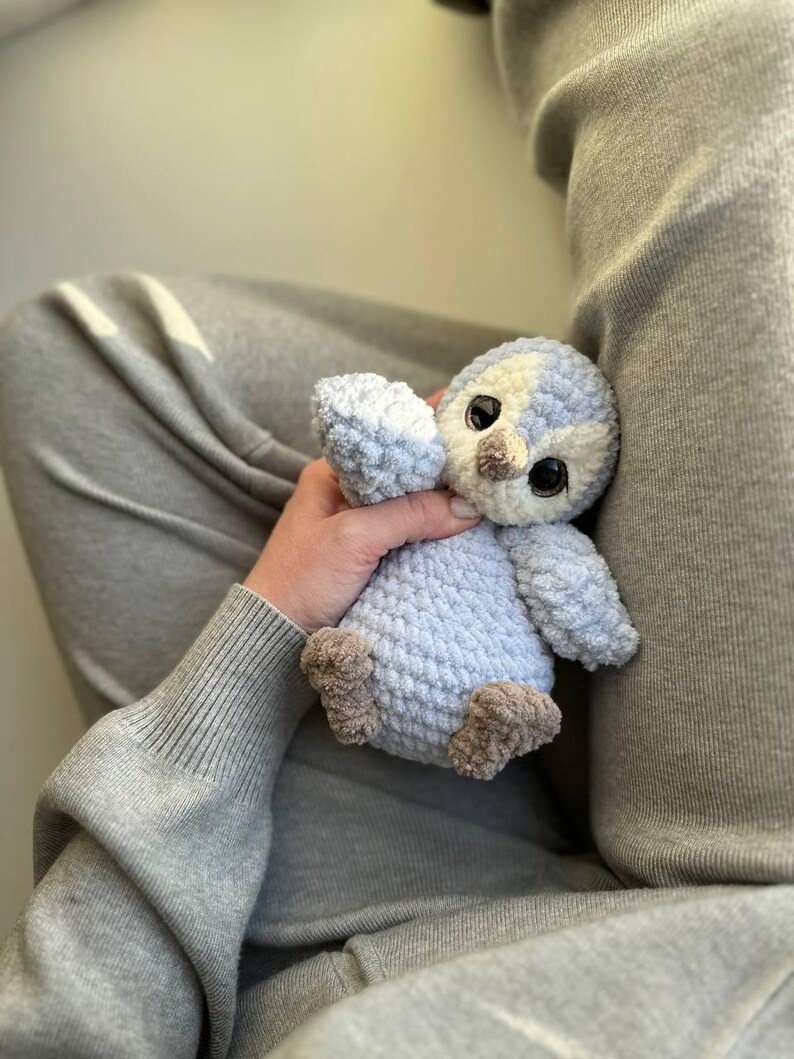 May include: A small, handmade crochet plush toy shaped like a penguin. The bird features a soft, light blue and cream-colored yarn body with a textured, fuzzy appearance. It has large, dark, glossy plastic eyes and a small, light brown yarn beak.