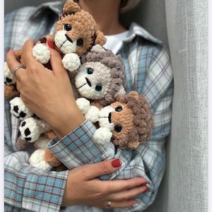 Könnte beinhalten: Drei gehäkelte Löwen- und Tigerbabys in Braun-, Beige- und Grautönen werden in den Armen gehalten. Die Babys haben weiße Pfoten und Gesichter mit schwarzen Augen und gestickten Details. Der Text "Crochet Pattern 'Mini Lion and Tiger Cub'" befindet sich unten.