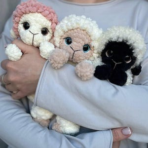 Puede incluir: Tres muñecos de oveja hechos a mano con ganchillo en una variedad de colores. Una oveja tiene la cabeza rosa, otra es blanca y la tercera es negra. Cada muñeco tiene ojos grandes y oscuros y un cuerpo esponjoso y texturizado. Los muñecos se sostienen juntos.