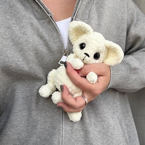 May include: A small, cream-colored crocheted puppy with large black eyes and floppy ears. The handmade stuffed animal is held in a person's hand, showcasing its soft texture and detailed craftsmanship. The puppy has a small red collar.