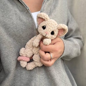 May include: A beige crocheted bunny plush toy with pink accents on the ears and feet. The bunny has large black eyes and a pink nose. The toy is held against a gray hooded sweatshirt.