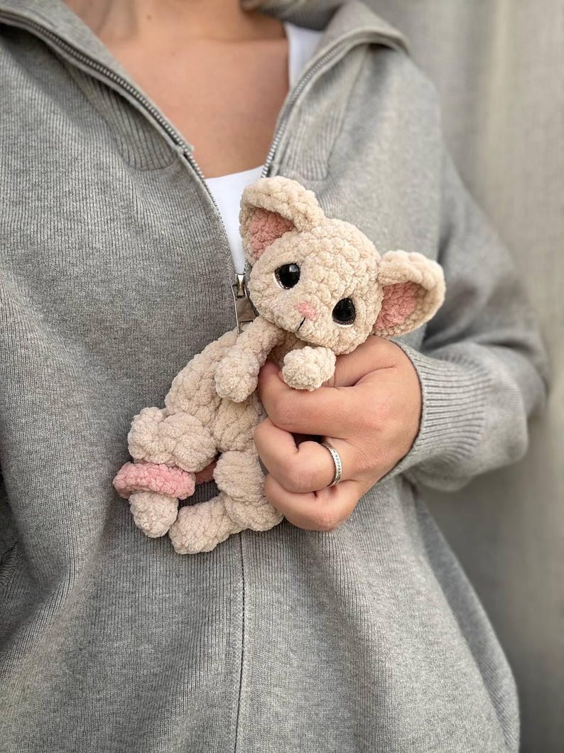 May include: A beige crocheted bunny plush toy with pink accents on the ears and feet. The bunny has large black eyes and a pink nose. The toy is held against a gray hooded sweatshirt.
