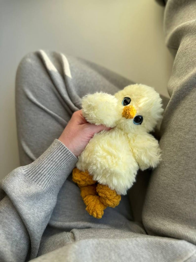 May include: A plush toy shaped like a small chick with soft, pale yellow fur, large dark eyes, a small orange beak, and textured orange feet. The stuffed animal has a rounded body and fluffy wings, crafted from synthetic materials.