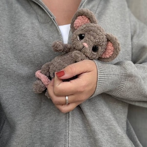 May include: A handmade, plush mouse toy in shades of gray and pink. The mouse has large, black eyes, pink inner ears, and a pink nose. The toy is held against a gray zip-up hoodie.
