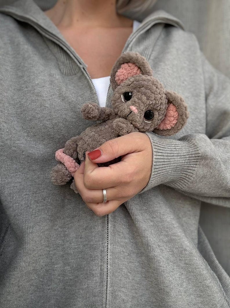 May include: A handmade, plush mouse toy in shades of gray and pink. The mouse has large, black eyes, pink inner ears, and a pink nose. The toy is held against a gray zip-up hoodie.