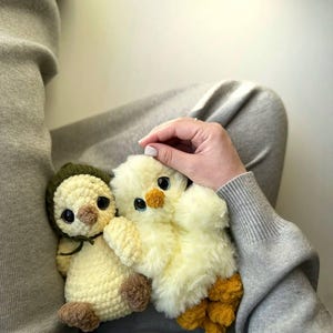May include: Two plush toys, one a cream-colored crocheted chick wearing a dark olive green bonnet with brown feet and beak, and the other a fluffy pale yellow chick with a small orange beak and textured orange feet, both featuring large dark eyes.