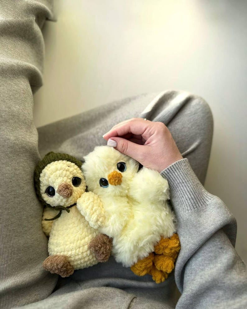 May include: Two plush toys, one a cream-colored crocheted chick wearing a dark olive green bonnet with brown feet and beak, and the other a fluffy pale yellow chick with a small orange beak and textured orange feet, both featuring large dark eyes.