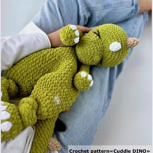 Puede incluir: Un juguete de dinosaurio de crochet verde con detalles blancos. El juguete está en el regazo de una persona. El texto "Crochet pattern<<Cuddle DINO>>" es visible en la parte inferior de la imagen.