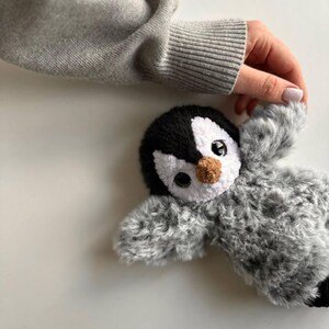 May include: A plush penguin toy featuring a black and white head with a small brown nose and large dark eyes. The body is covered in soft, textured gray and white faux fur, giving it a fuzzy appearance. The toy has outstretched wings.