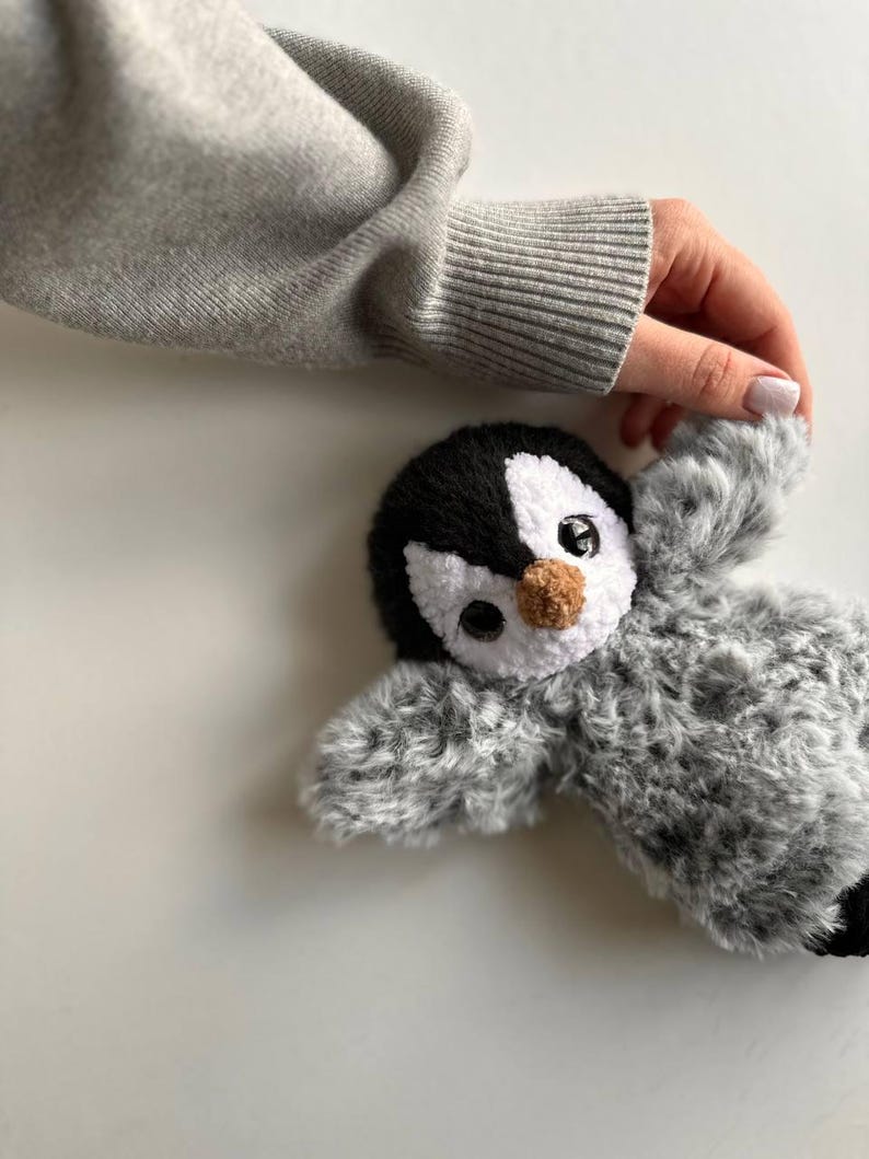 May include: A plush penguin toy featuring a black and white head with a small brown nose and large dark eyes. The body is covered in soft, textured gray and white faux fur, giving it a fuzzy appearance. The toy has outstretched wings.